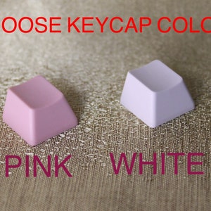 May include: Two square keycaps, one pink and one white, are displayed against a textured gold background. The text "CHOOSE KEYCAP COLOR" is above the keycaps, and "PINK WHITE" is below them.