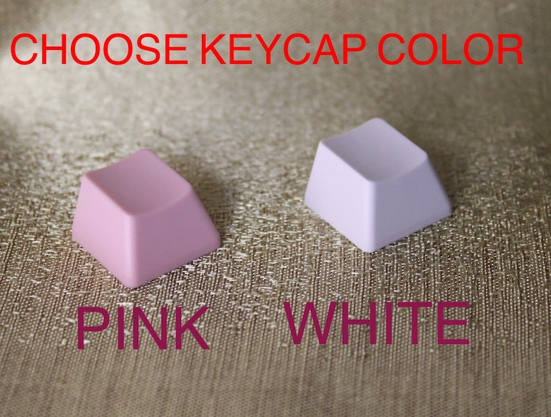 May include: Two square keycaps, one pink and one white, are displayed against a textured gold background. The text "CHOOSE KEYCAP COLOR" is above the keycaps, and "PINK WHITE" is below them.