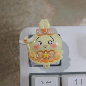 May include: A yellow, cartoon-style keyboard keycap featuring a smiling character with pink cheeks, a bow, and a star. The keycap has a wand and is set against a white keyboard.