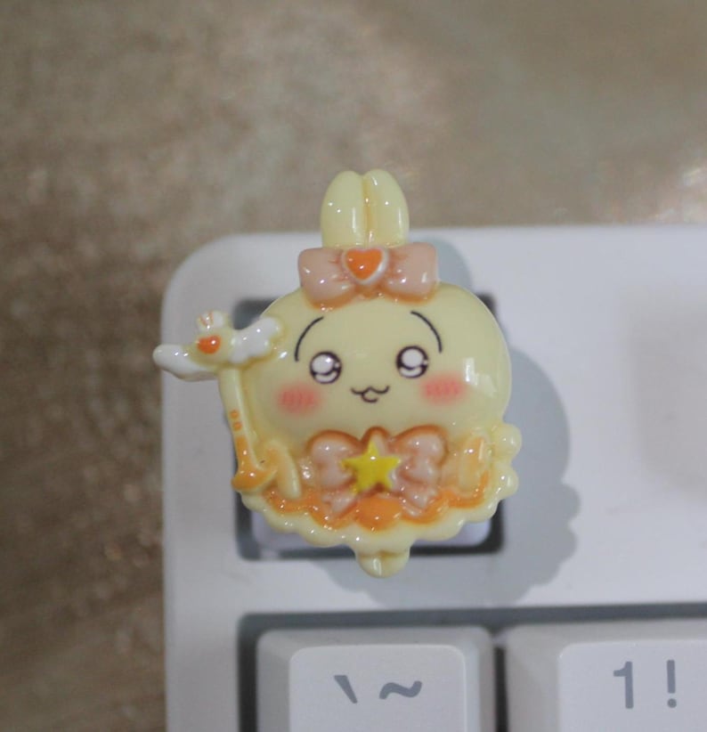 May include: A yellow, cartoon-style keyboard keycap featuring a smiling character with pink cheeks, a bow, and a star. The keycap has a wand and is set against a white keyboard.