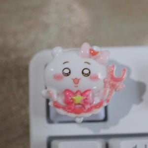 May include: A white and pink cartoon character keycap with a bow and star design. The keycap is on a white keyboard. The character has large eyes and a smiling expression. The keycap is a decorative accessory for a mechanical keyboard.