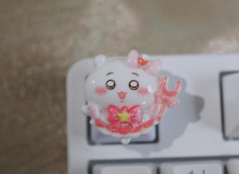May include: A white and pink cartoon character keycap with a bow and star design. The keycap is on a white keyboard. The character has large eyes and a smiling expression. The keycap is a decorative accessory for a mechanical keyboard.