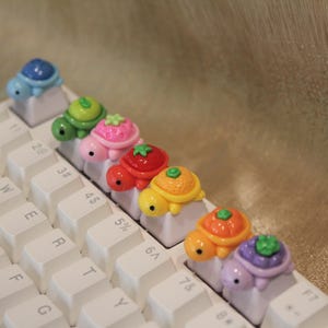Puede incluir: Un teclado de computadora blanco con una fila de teclas personalizadas, cada una rematada con una pequeña figura de tortuga colorida. Las tortugas tienen caparazones con motivos de frutas, incluyendo diseños de fresa, tomate, naranja y uva.