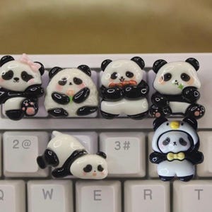 May include: A collection of seven panda-shaped keycaps in various poses. The keycaps are primarily white and black, with pink accents on some. One panda wears a bow, another a hat with a yellow bow tie. They are placed on a white keyboard.