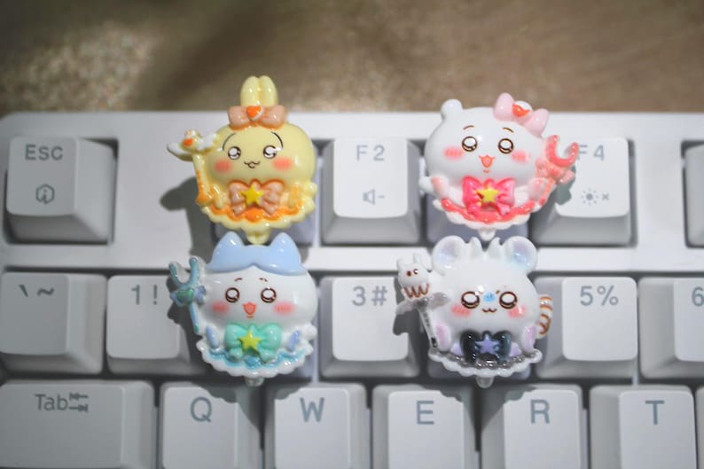 May include: Four colorful, cartoon-style keycaps with cute character designs are arranged on a white keyboard. The keycaps feature characters in yellow, pink, blue, and white, with various accessories and expressions. The keyboard keys are white.