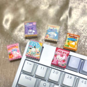 Könnte beinhalten: Eine Sammlung farbenfroher Miniatur-Anhänger in Form von Süßigkeitenbeuteln mit Zeichentrickfiguren und japanischem Text. Die Anhänger sind auf einer Computertastatur angeordnet. Die Beutel sind rosa, blau, lila, gelb und orange.