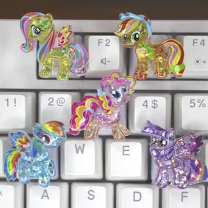 May include: Five colorful glittery pony figurines on a white keyboard. The ponies are all different colors and have different designs. The keyboard has the keys 1!, 2@, 4$, 5%, W, E, T, A, S, D, F.