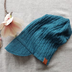 Könnte beinhalten: Eine türkisfarbene Strickmütze mit einem Schirm und einem braunen Lederlabel. Die Mütze hat ein Zopfmuster.