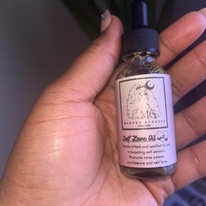 May include: A clear glass bottle with a black dropper top and a pink label that reads "Self Love Oil". The label also includes the text "Handcrafted and spelled to aid in boosting self esteem. Promote inner peace, confidence and self love."