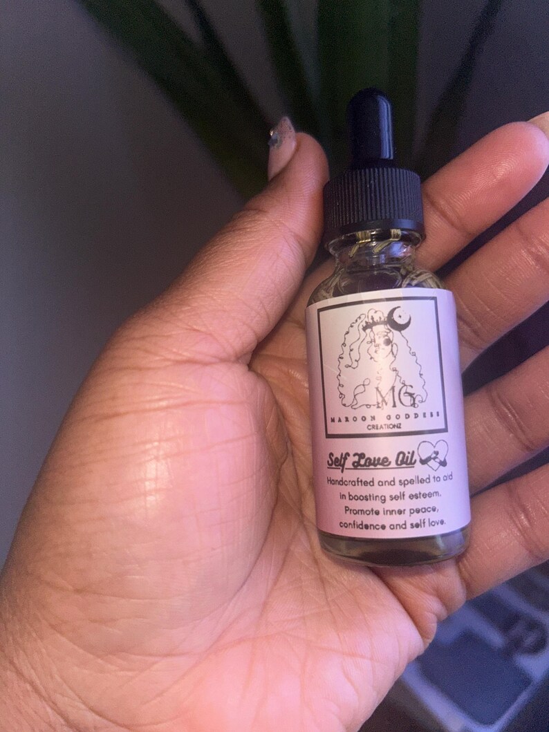 May include: A clear glass bottle with a black dropper top and a pink label that reads "Self Love Oil". The label also includes the text "Handcrafted and spelled to aid in boosting self esteem. Promote inner peace, confidence and self love."