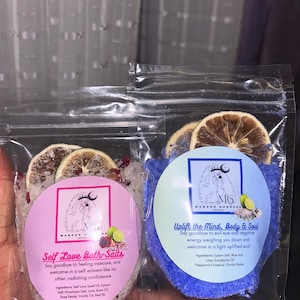 May include: Two small bags of bath salts with dried citrus fruit on top. The bag on the left is pink and says "Self Love Bath Salts" with a list of ingredients. The bag on the right is blue and says "Uplift the Mind, Body & Soul" with a list of ingredients.