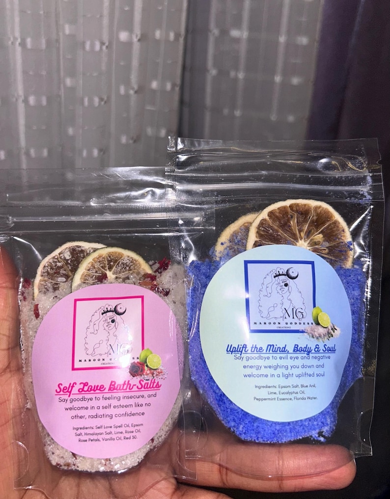 May include: Two small bags of bath salts with dried citrus fruit on top. The bag on the left is pink and says "Self Love Bath Salts" with a list of ingredients. The bag on the right is blue and says "Uplift the Mind, Body & Soul" with a list of ingredients.