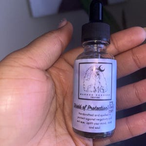 May include: A clear glass bottle with a black dropper top and a white label. The label has a black and white illustration of a woman with a crown and the text "MG", "Magick Goddess Creations", and "Shield of Protection". The text on the label reads "Handcrafted and spelled to protect against negativity and evil eye. Uplift your mind, body and soul."
