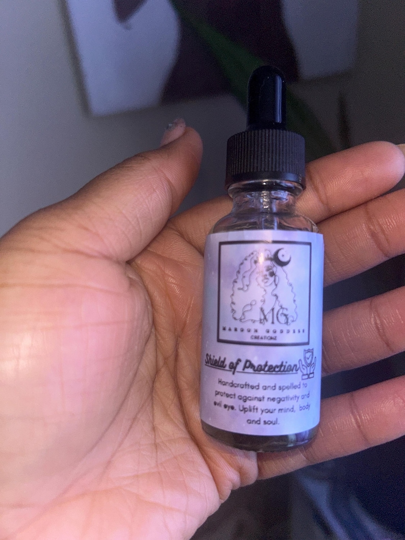May include: A clear glass bottle with a black dropper top and a white label. The label has a black and white illustration of a woman with a crown and the text "MG", "Magick Goddess Creations", and "Shield of Protection". The text on the label reads "Handcrafted and spelled to protect against negativity and evil eye. Uplift your mind, body and soul."