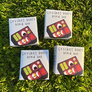 May include: Four small square boxes with a bento box design are displayed on a bed of green grass. Each box features the text "Crochet bento blind box" and a colorful illustration of a bento box with sushi and other food items.