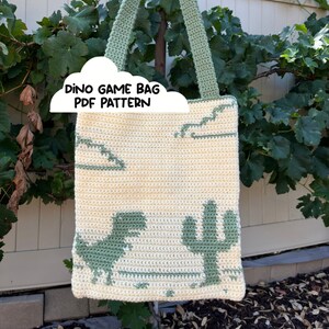 Op de afbeelding: Een gehaakte tote bag met een ontwerp van een groene dinosaurus, cactus en wolken. De tas heeft een groene handvat en is gemaakt van crème en groen garen. De tekst "Dino Game Bag PDF Pattern" staat op de tas.