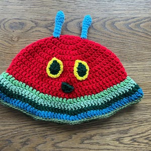 May include: A handmade crochet caterpillar hat. The hat is predominantly red with a green, blue, and yellow striped brim. It has yellow eyes, a green nose, and blue antennae. A fun, colourful accessory, perfect for children.