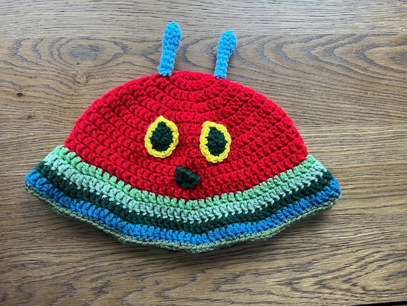 May include: A handmade crochet caterpillar hat. The hat is predominantly red with a green, blue, and yellow striped brim. It has yellow eyes, a green nose, and blue antennae. A fun, colourful accessory, perfect for children.
