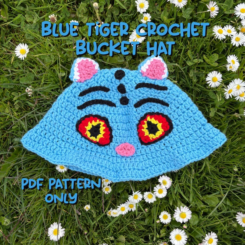 Puede incluir: Un sombrero de pescador de ganchillo azul claro con un dise&ntilde;o de cara de tigre. El sombrero presenta orejas rosas, detalles negros y ojos grandes con acentos rojos y amarillos. El texto "BLUE TIGER CROCHET BUCKET HAT" y "PDF PATTERN ONLY" son visibles.