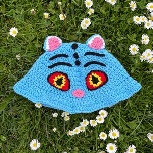Puede incluir: Un sombrero de ganchillo azul claro hecho a mano con un dise&ntilde;o de cara de gato. El sombrero presenta orejas rosas, cejas negras y ojos grandes con detalles rojos y amarillos. El sombrero est&aacute; sobre hierba verde con margaritas blancas.