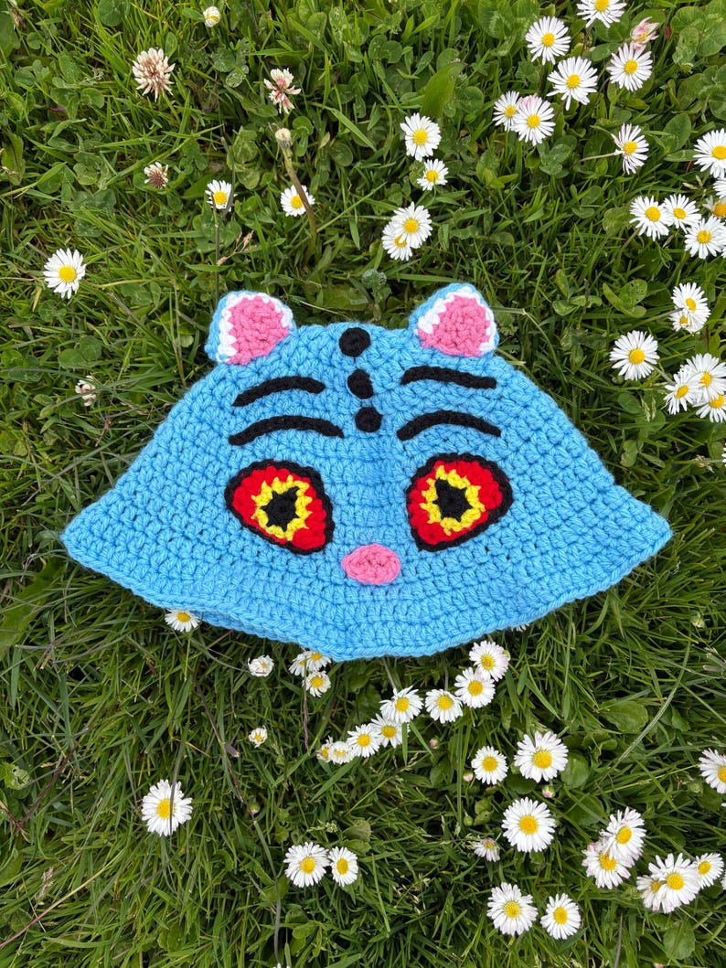 Puede incluir: Un sombrero de ganchillo azul claro hecho a mano con un dise&ntilde;o de cara de gato. El sombrero presenta orejas rosas, cejas negras y ojos grandes con detalles rojos y amarillos. El sombrero est&aacute; sobre hierba verde con margaritas blancas.