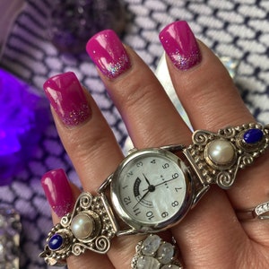 May include: A silver watch with a white face and black numbers. The watch has a decorative band with pearls and blue stones. The watch is on a person's wrist.