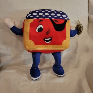 Op de afbeelding: Een rood pluche speelgoed in de vorm van een boek met een blauw-witte stippenhoed, een zwart ooglapje en een geel frame. Het speelgoed heeft blauwe benen en witte schoenen. Het speelgoed houdt een klein bruin en gouden voorwerp in zijn rechterhand.