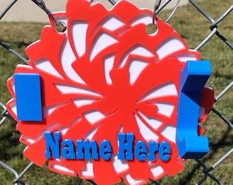 May include: A circular red and white cheerleading pom-pom shaped sign with two blue hooks attached to the sides. The sign features the blue text Name Here in the center and is suspended by two metal rings from a chain-link fence.