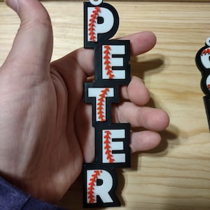Puede incluir: Un adorno de béisbol en blanco y negro que deletrea "PETER" con detalles de costura rojos. Las letras están apiladas verticalmente, con un pequeño agujero en la parte superior para colgar. El adorno se sostiene en una mano.