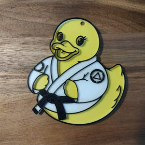Op de afbeelding: Een gele eend met een witte karate gi met een zwarte band. De eend heeft zwarte ogen en een zwarte omtrek. De gi heeft een patch met een driehoek symbool. De eend is een sleutelhanger of ornament.