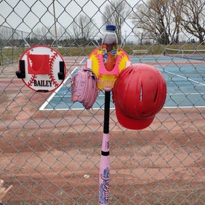 May include: A baseball-themed setup with a pink bat, a red helmet, and a pink glove hanging on a chain-link fence. A bottle of juice sits in a holder. A sign with the name "Bailey" is visible.
