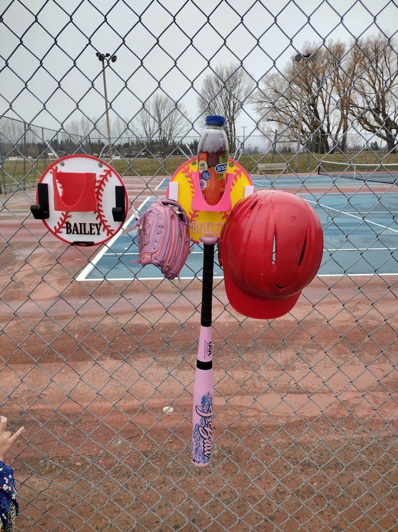 May include: A baseball-themed setup with a pink bat, a red helmet, and a pink glove hanging on a chain-link fence. A bottle of juice sits in a holder. A sign with the name "Bailey" is visible.