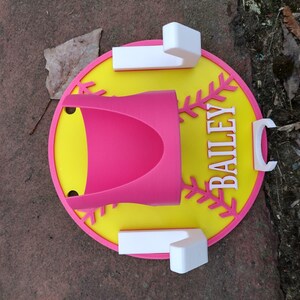May include: A pink and yellow baseball-themed holder with the name "BAILEY" in white. The holder has a pink cup-shaped section and white rectangular and C-shaped accents. The design is set on a brown surface.