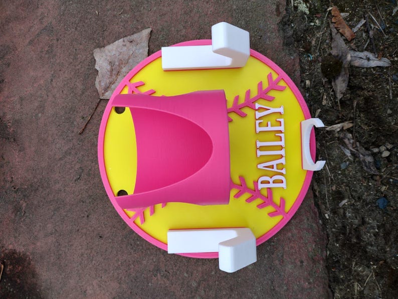 May include: A pink and yellow baseball-themed holder with the name "BAILEY" in white. The holder has a pink cup-shaped section and white rectangular and C-shaped accents. The design is set on a brown surface.