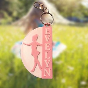 May include: A pink and white keychain featuring a ballerina silhouette and the name "EVELYN" in capital letters. The keychain has a silver-colored clasp and ring. The background is a blurred outdoor scene with green grass and sunlight.