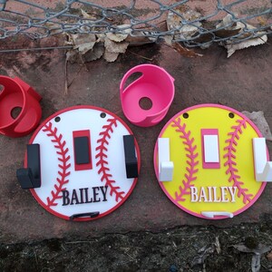 May include: Baseball-themed wall hooks and holders in red, white, pink, and yellow. The hooks are round with a baseball stitch design and the name "BAILEY". Two red and pink holders are also included.