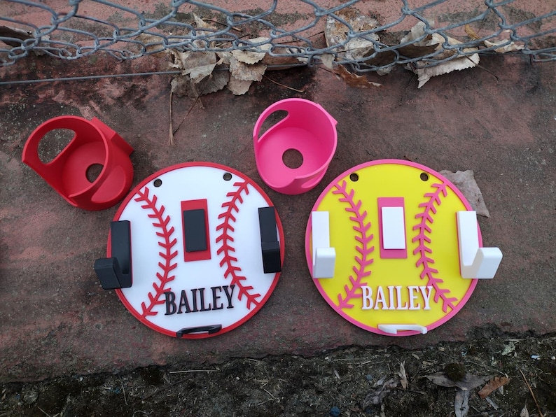 May include: Baseball-themed wall hooks and holders in red, white, pink, and yellow. The hooks are round with a baseball stitch design and the name "BAILEY". Two red and pink holders are also included.