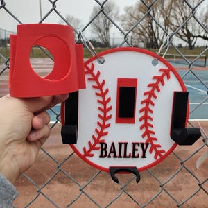 May include: A red and white baseball-themed holder with the name "BAILEY" in black. The holder has a circular design with red stitching and a red border. It includes a red cup holder and black hooks.