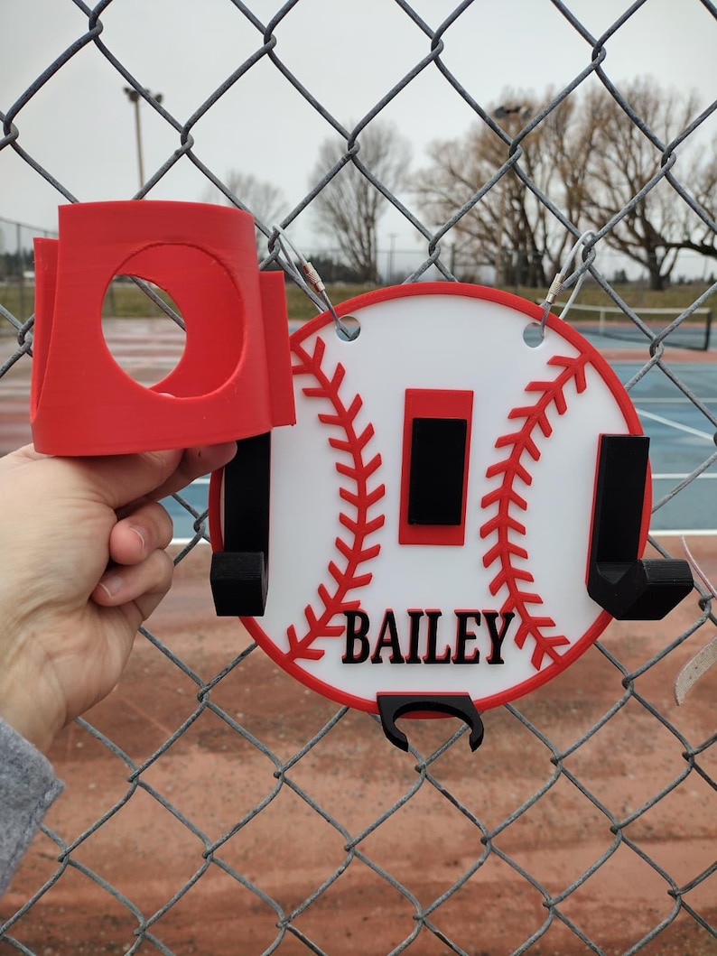 May include: A red and white baseball-themed holder with the name "BAILEY" in black. The holder has a circular design with red stitching and a red border. It includes a red cup holder and black hooks.