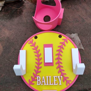 May include: A softball-themed wall organizer in yellow, pink, and white. The design includes a yellow circle with pink stitching and trim, white hooks, and the name "BAILEY". A pink holder is also included.