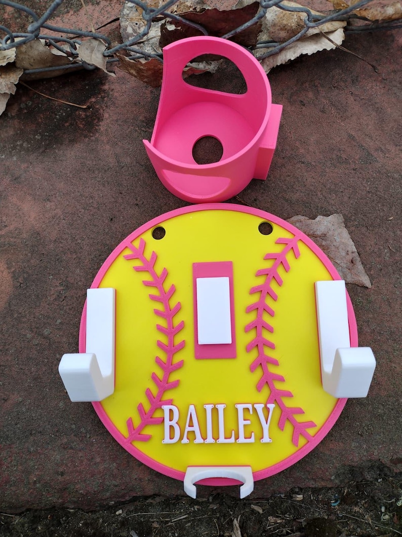 May include: A softball-themed wall organizer in yellow, pink, and white. The design includes a yellow circle with pink stitching and trim, white hooks, and the name "BAILEY". A pink holder is also included.