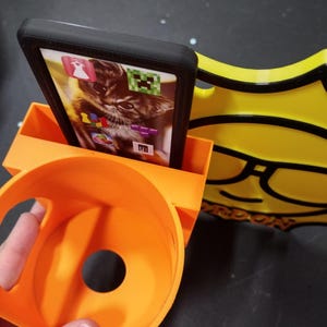 May include: An orange 3D-printed phone stand with a built-in cup holder and a yellow decorative element. A black phone case holds a phone displaying a cat image and app icons. The stand is designed to hold a phone upright.