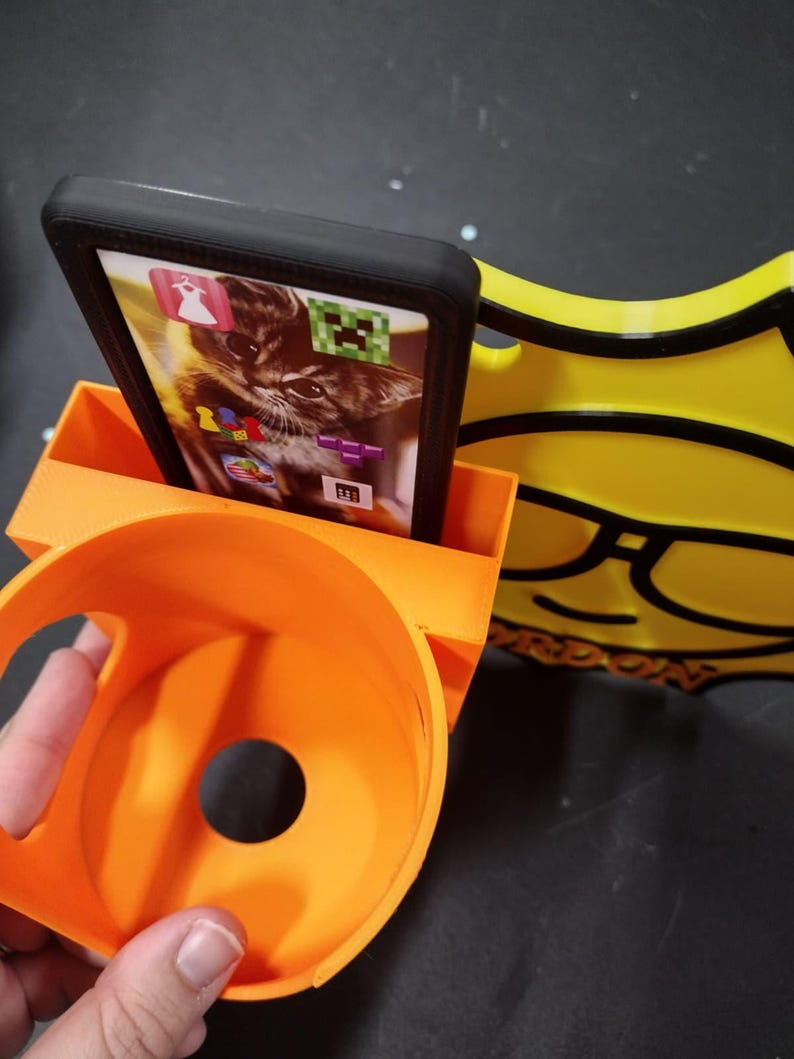 May include: An orange 3D-printed phone stand with a built-in cup holder and a yellow decorative element. A black phone case holds a phone displaying a cat image and app icons. The stand is designed to hold a phone upright.