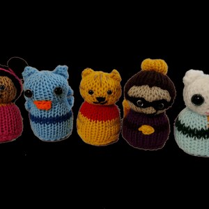 May include: Five knitted plush toys, each with a different animal design. The toys are all in a row, from left to right: a brown girl with a pink dress, a blue bird with a yellow beak, a yellow cat with a red shirt, a brown sloth with a purple shirt, and a white cat with a blue and green shirt.
