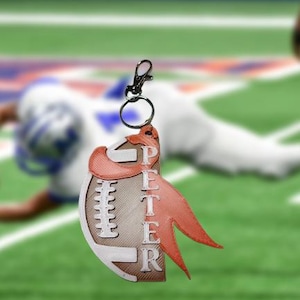 May include: A football-shaped keychain with the name "PETER" in white letters. The keychain has a brown and white football design with a red ribbon-like accent and a silver clasp.