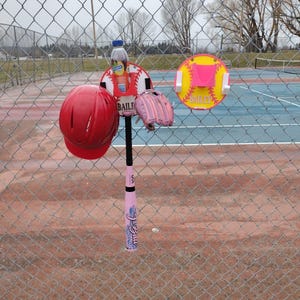 May include: A red softball helmet, pink softball glove, and a pink softball bat are displayed on a chain-link fence. A softball-themed sign with the name "Bailey" is also visible. A bottle of juice is attached to the softball sign.