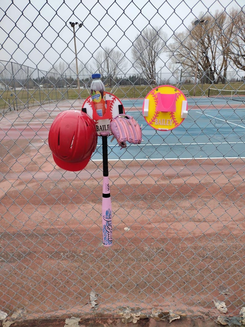 May include: A red softball helmet, pink softball glove, and a pink softball bat are displayed on a chain-link fence. A softball-themed sign with the name "Bailey" is also visible. A bottle of juice is attached to the softball sign.