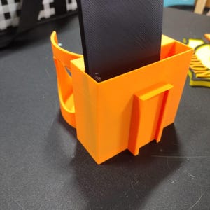 May include: Bright orange 3D-printed phone holder with a handle and a rectangular compartment holding a black phone. The holder has a square shape with a small rectangular extension on the side.