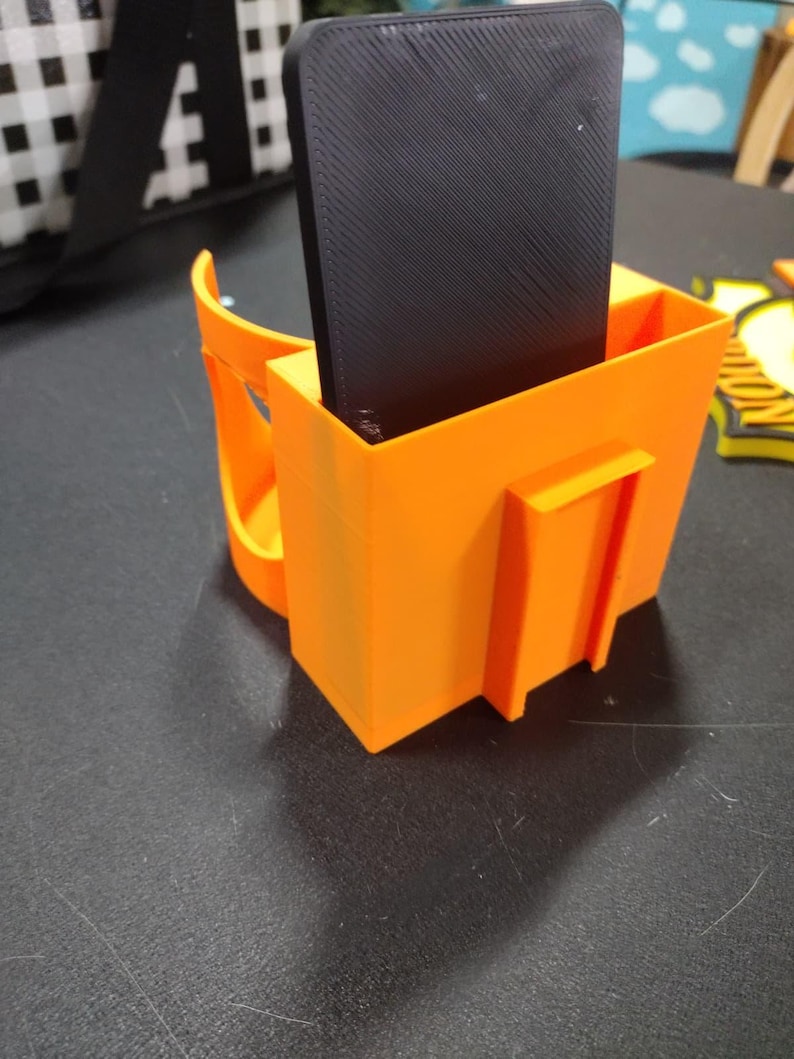 May include: Bright orange 3D-printed phone holder with a handle and a rectangular compartment holding a black phone. The holder has a square shape with a small rectangular extension on the side.
