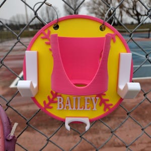 May include: A personalised softball holder in yellow and pink. The holder has the name "BAILEY" in white lettering. It features a pink centre and white hooks for hanging. The holder is attached to a chain-link fence.